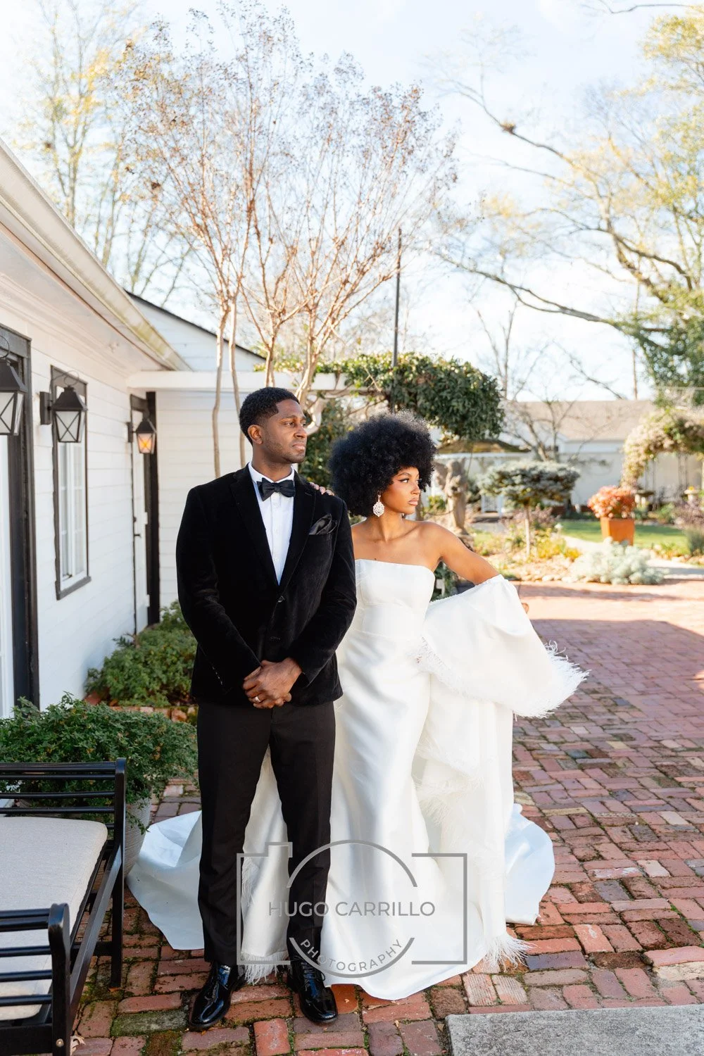 A couple dressed in formal attire, with the man in a black tuxedo and the woman in a white wedding gown, standing outdoors on a brick pathway near a white house with greenery and trees in the background.