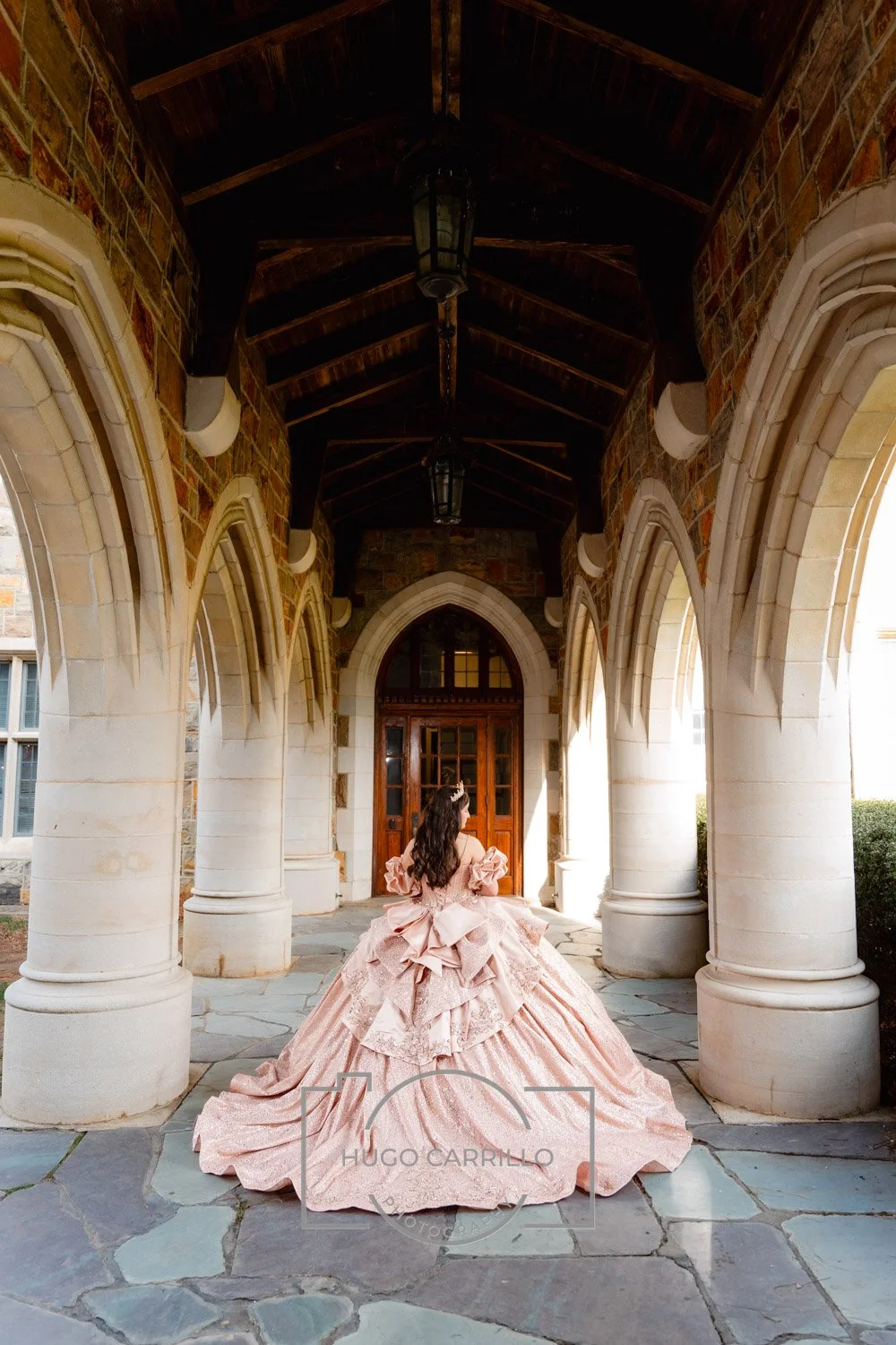 A quinceañera in an elaborate pink ball gown with ruffles and bows, standing under a stone and brick archway with wooden beams and lanterns, facing away from the camera.