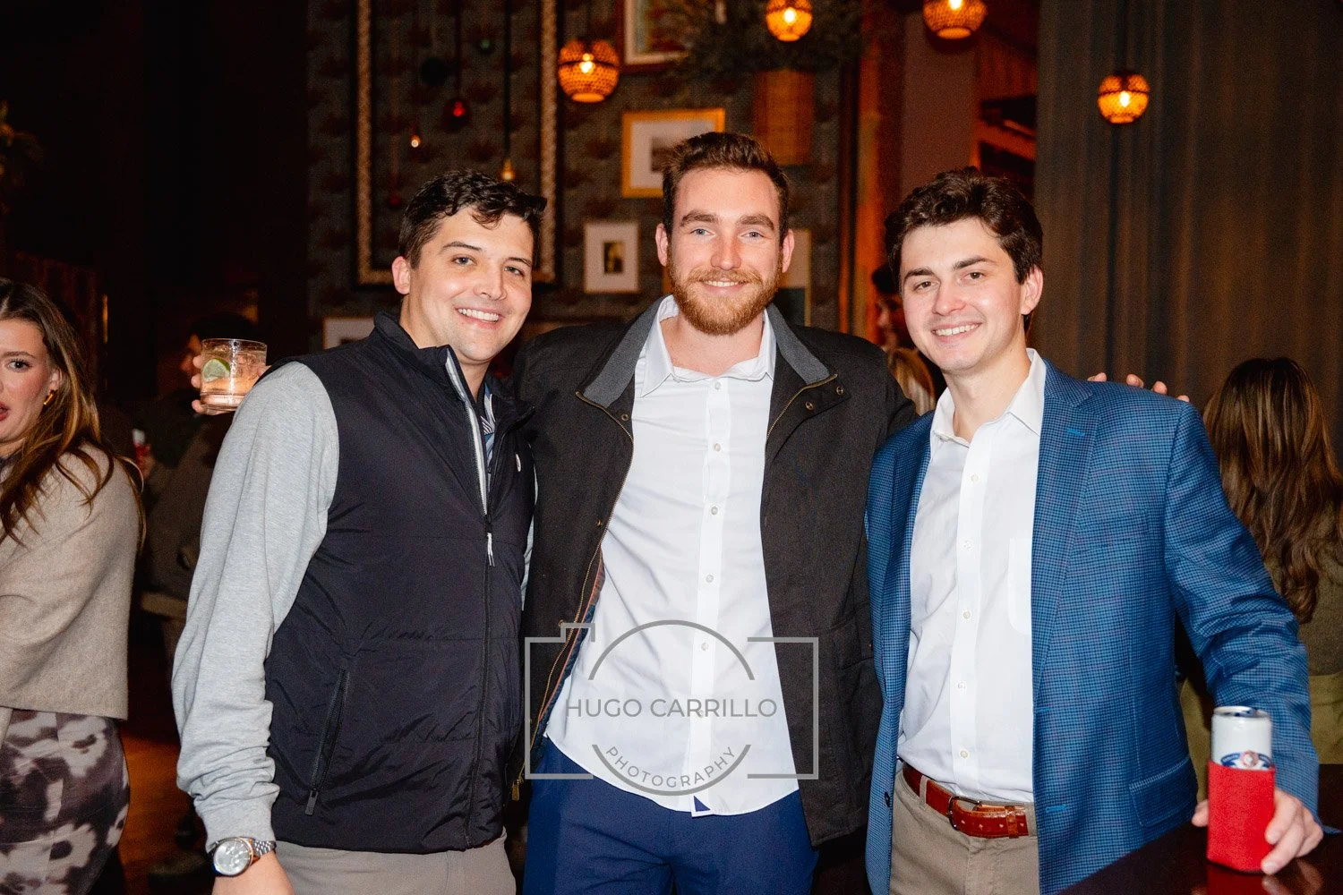 Three young men smiling and posing at a social gathering. The man on the left is wearing a black and gray jacket, the man in the middle is wearing a black jacket over a white shirt, and the man on the right is wearing a blue suit jacket with a white 