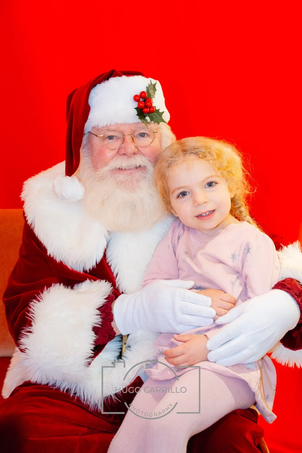 Santa Claus with a white beard and glasses, wearing a red suit with white faux fur trim, sitting with a young girl with curly blonde hair and a pink dress, against a red background.