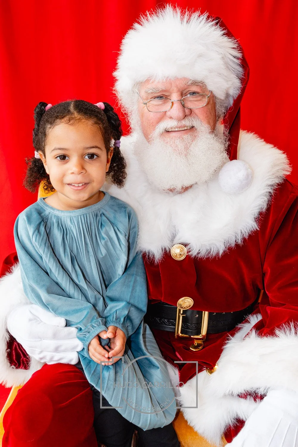 A young girl with curly hair and a blue dress sitting on Santa Claus's lap, who is dressed in a red suit with white fur trim, against a red background.