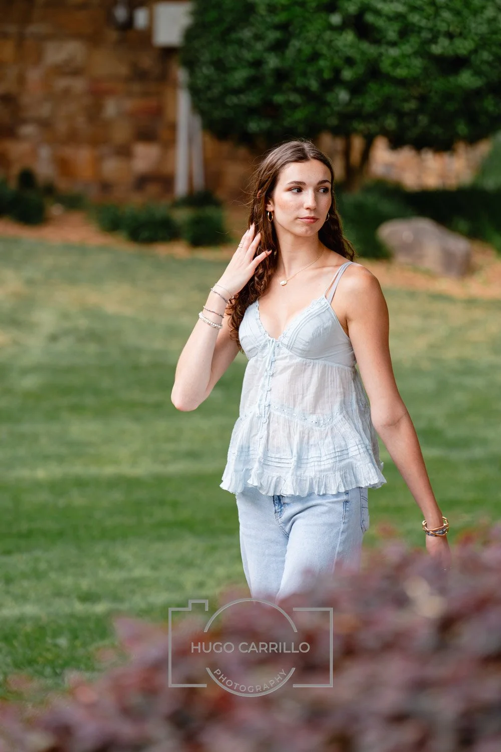 Young woman with long wavy hair wearing a white sleeveless top and light blue jeans, standing outdoors in a park-like setting with greenery and a building in the background.