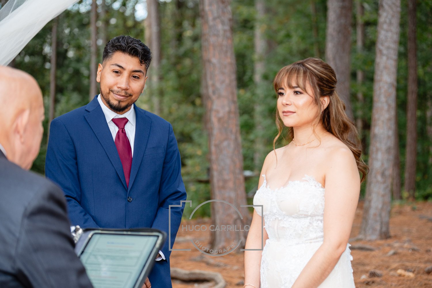 A couple of people standing outdoors in a forest during a wedding ceremony, with an officiant holding a tablet in front of them.