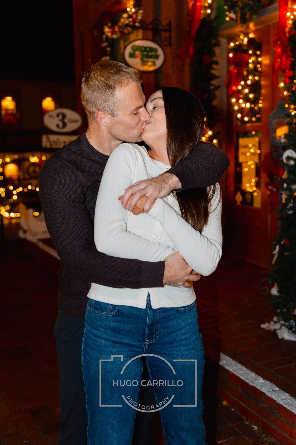 A couple sharing a kiss and hugging inside a warmly decorated space with Christmas lights and decorations.