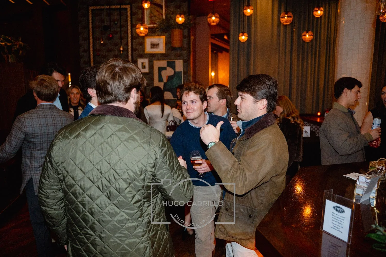 Group of people at a social gathering, chatting and holding drinks, in a warmly lit indoor setting with artwork and hanging lights on the walls.