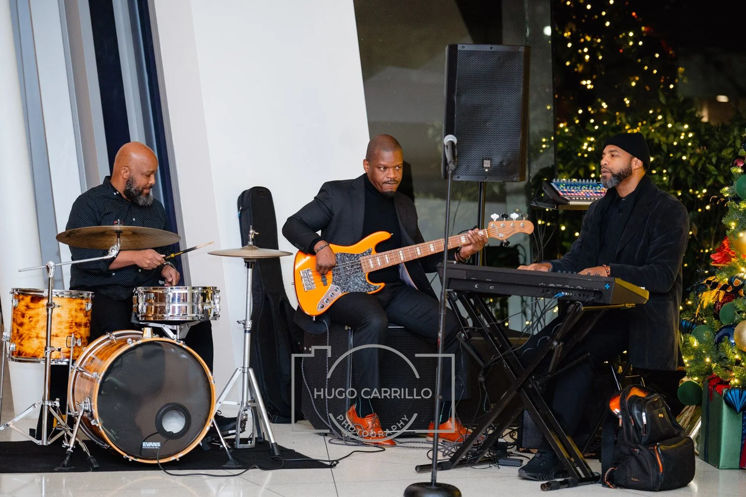 Three musicians playing instruments near a decorated Christmas tree, with one on drums, one on bass guitar, and one on keyboard, in an indoor setting.