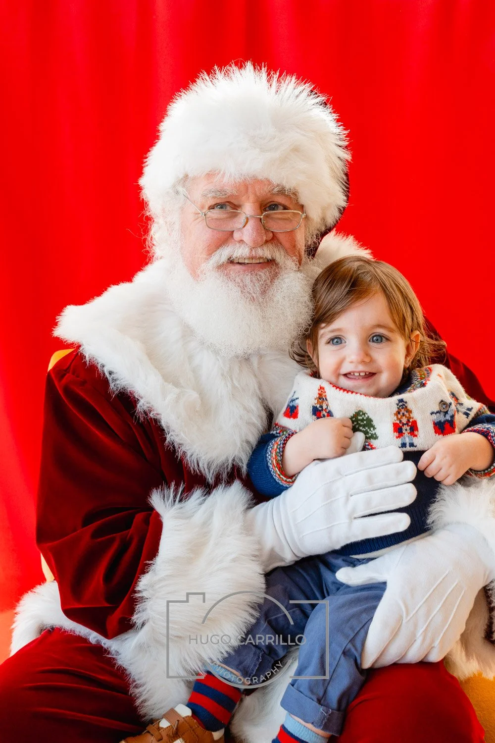 Santa Claus holding a young boy in front of a red background, smiling.