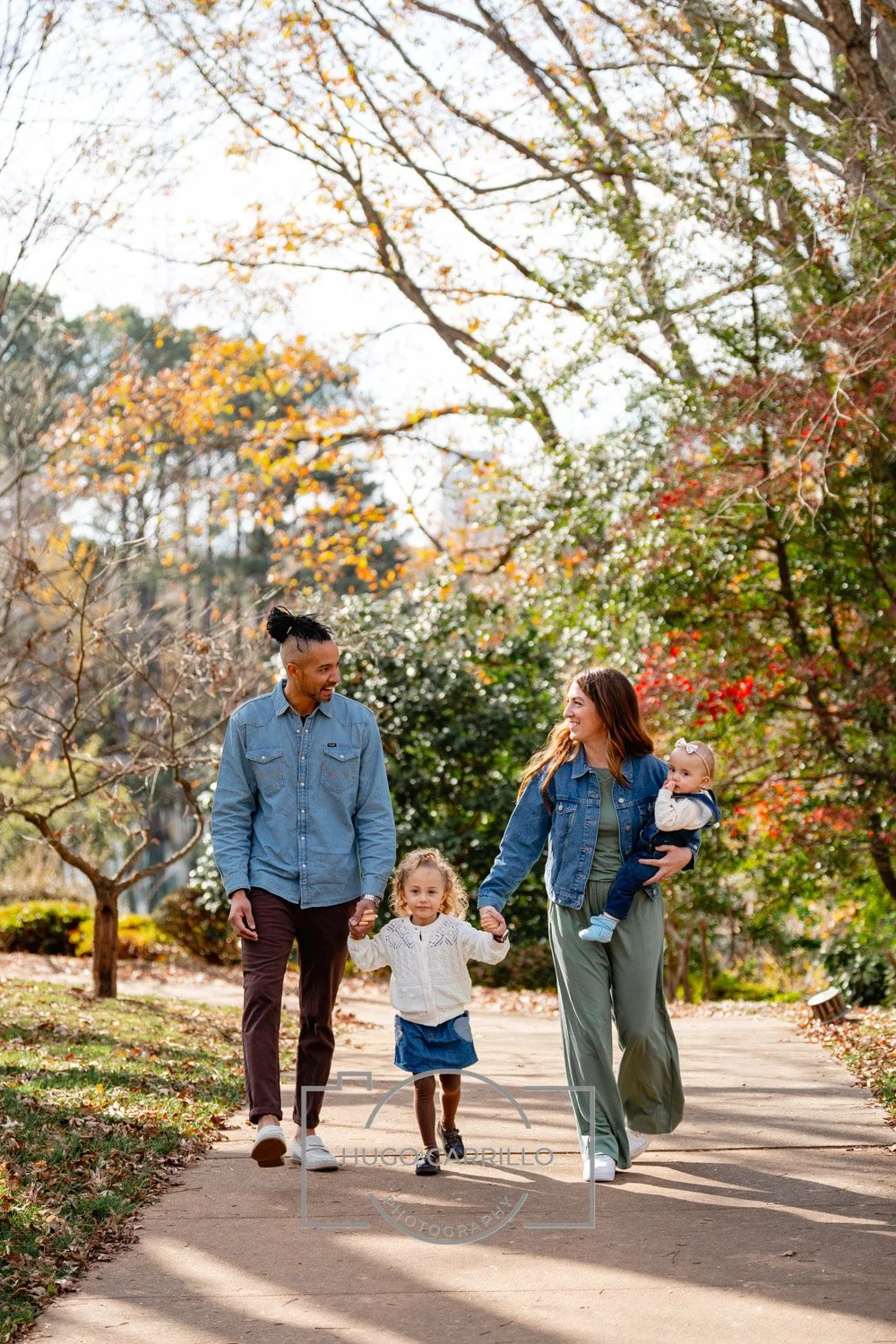 A family of four walking on a park path during fall with colorful trees in the background. The father and daughter hold hands, and the mother carries a baby.