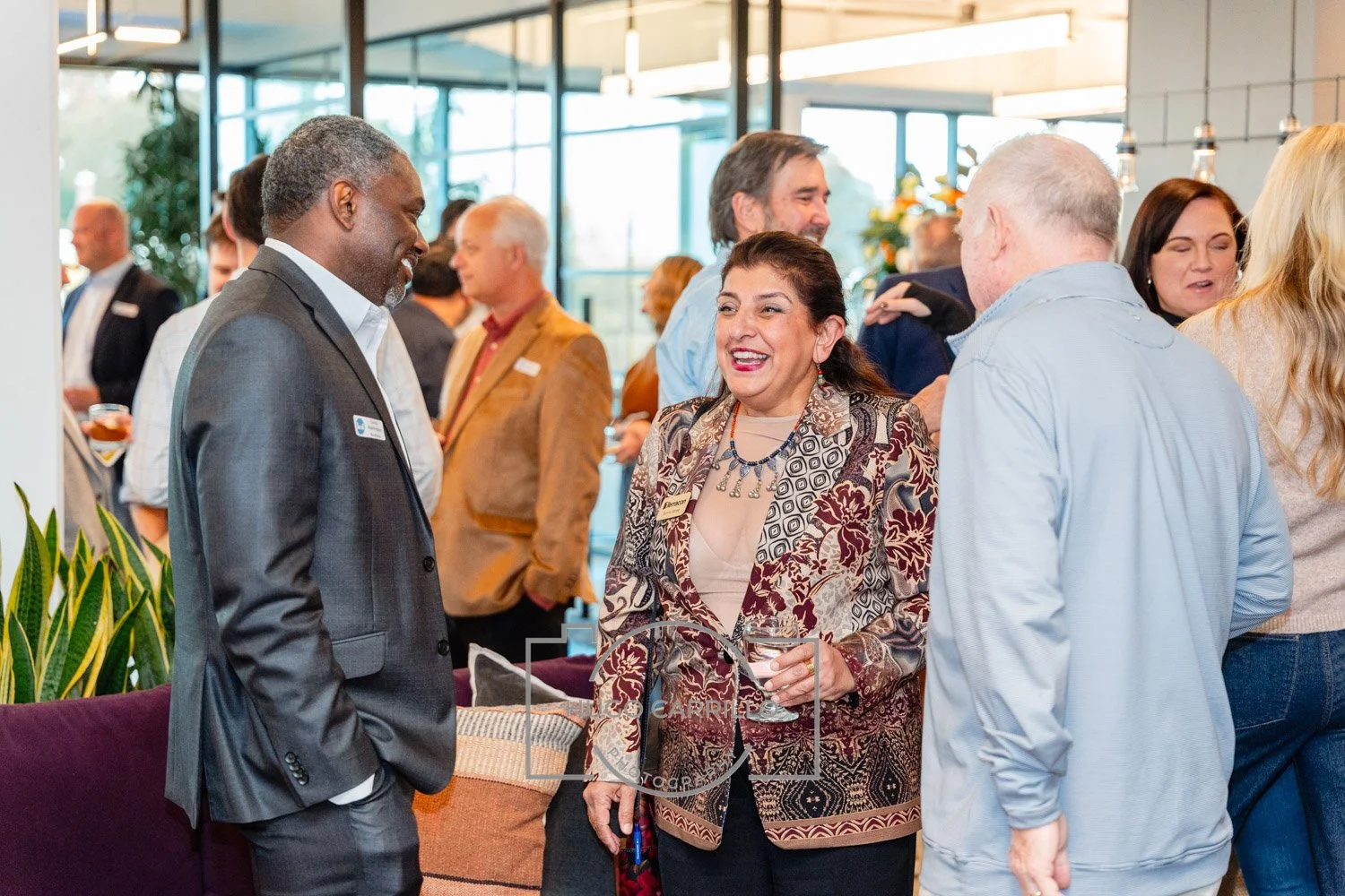 A group of people at a networking event. A woman with dark hair and a patterned blazer is smiling and holding a wine glass, talking to a man in a gray suit. Other attendees are mingling in the background.