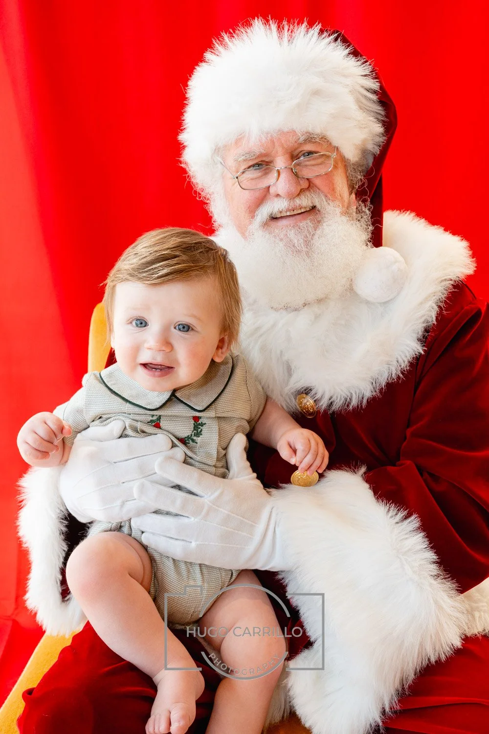 A young child sitting on Santa Claus's lap, with a red background, Santa wearing a red suit with white fur trim, glasses, and a white beard, and the child smiling and reaching out with one hand.