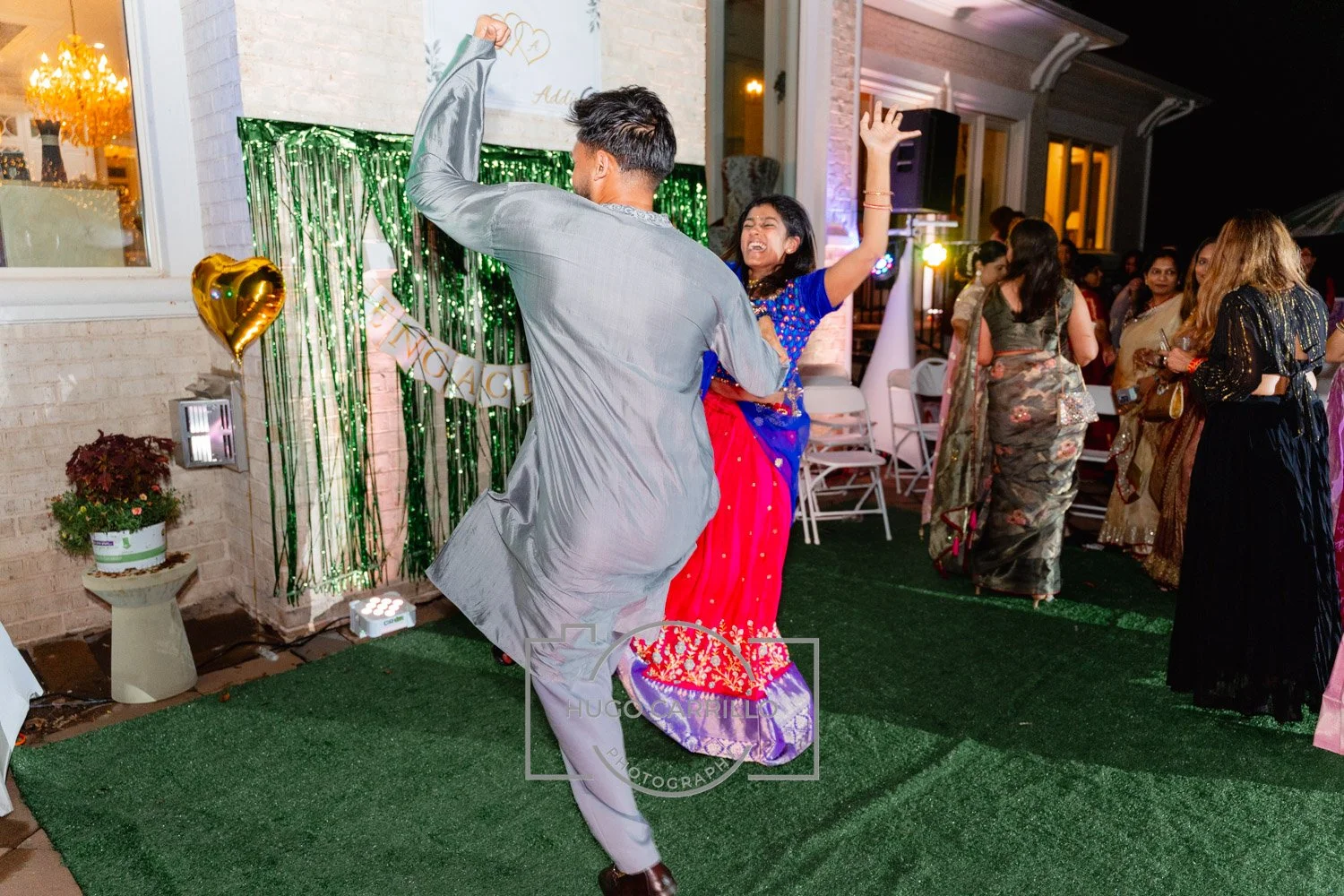 A man and a woman dancing happily at a celebration, with other women in traditional dresses in the background, and decorations including green streamers, gold balloons, and a banner reading "Congratulations."