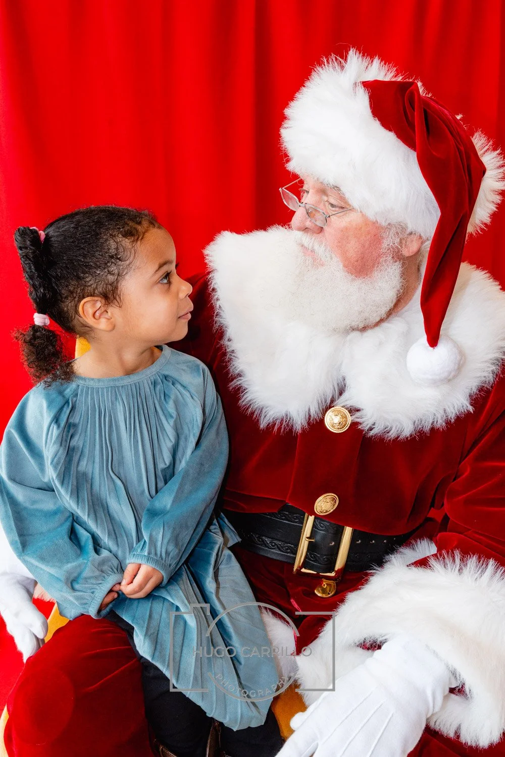 A young girl with curly hair looking at Santa Claus, who is dressed in a red velvet suit with white fur trim and a matching hat, sitting in front of a red curtain.