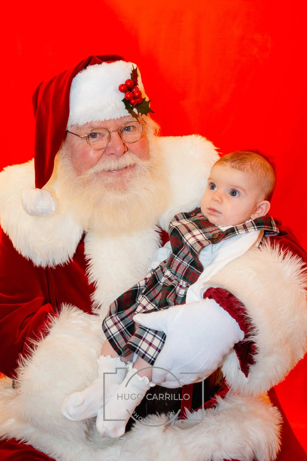 Santa Claus in a red suit with white fur trim and holly berries on his hat holding a young child in a plaid outfit against a red background.