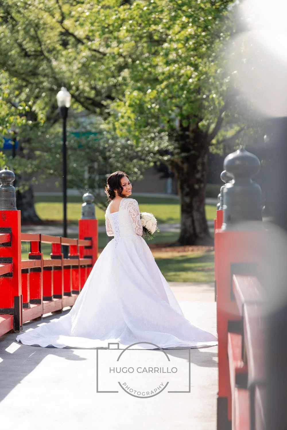 Bride standing on a bridge outdoors, wearing a white wedding gown and holding a bouquet, with trees and a lamppost in the background.