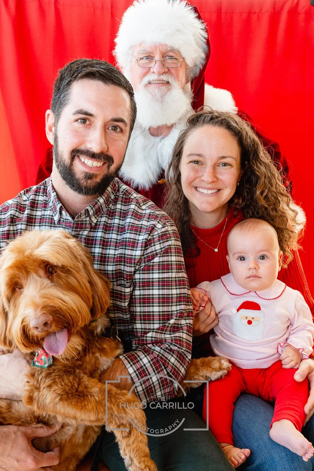 A family of three, a man, woman, and baby girl, sitting with a large brown dog, with Santa Claus standing behind them. The background is red, and everyone is smiling, dressed in festive clothing.