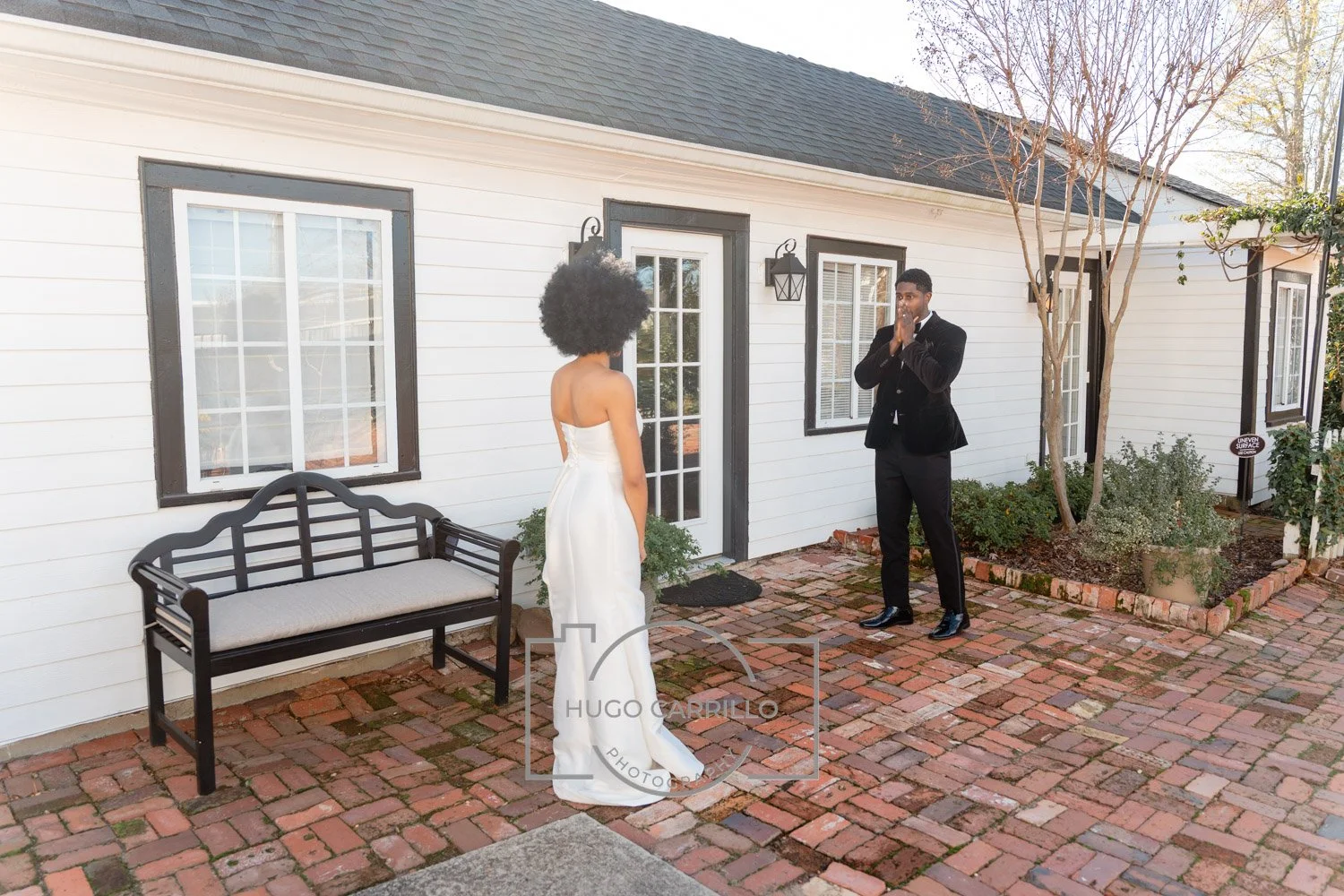 A woman in a white dress facing a man in a black suit outside a house, with the man appearing surprised or emotional. A black bench is on the brick patio beside the house.