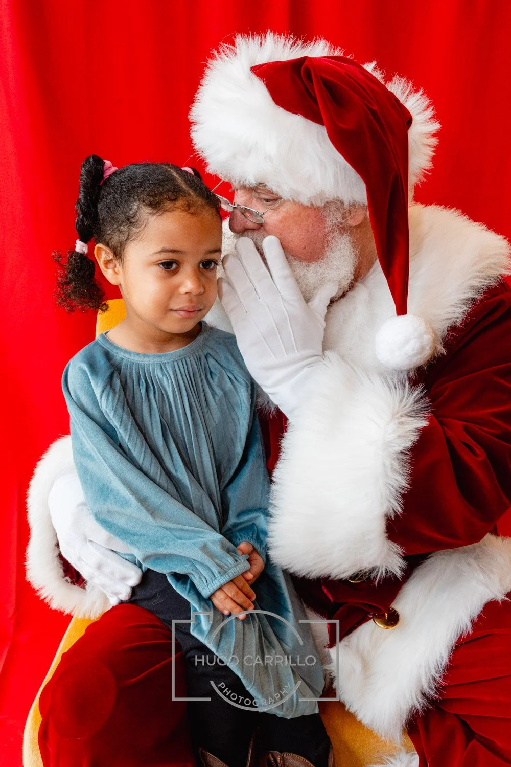 Santa Claus whispering to a young girl during a Christmas event, with a red curtain background.