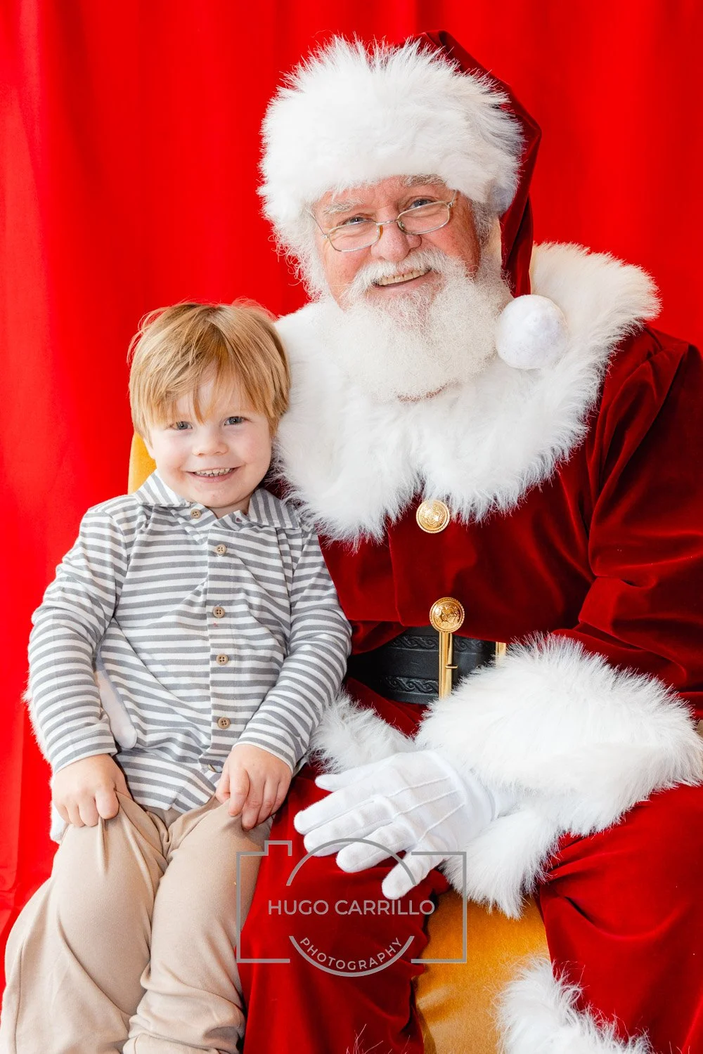 A young boy with red hair sitting on Santa Claus's lap, smiling, in front of a red curtain.