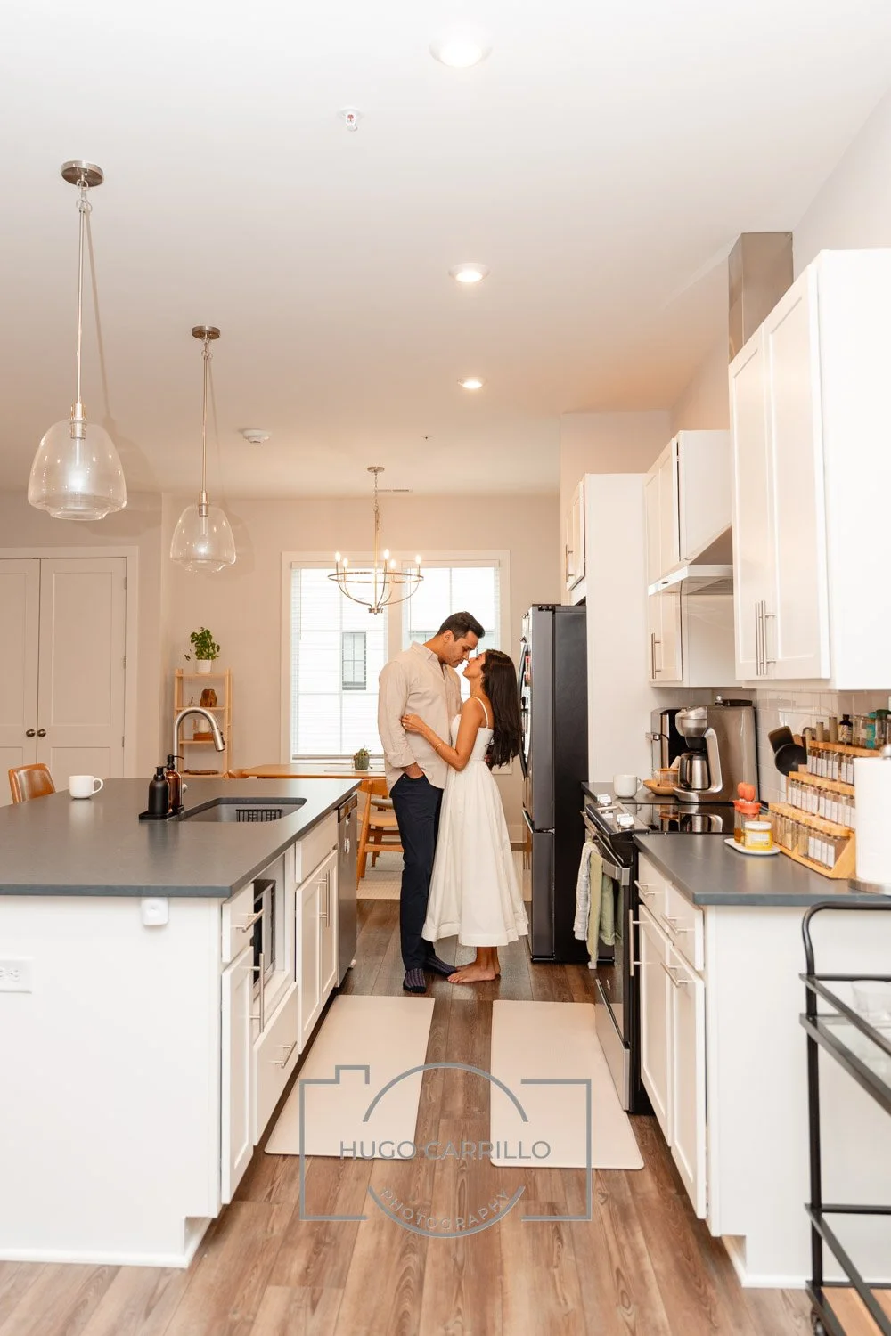 A man and woman standing close together in a kitchen, smiling and touching foreheads, with the woman in a white dress and the man in a light shirt and dark pants.