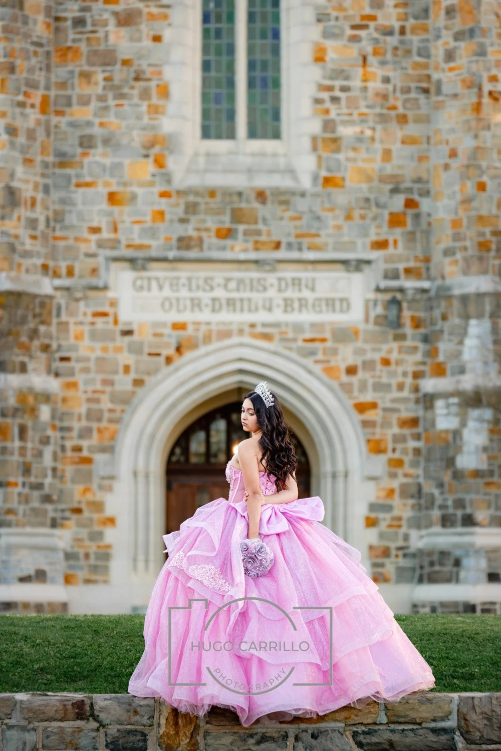 A quinceañera in a pink ball gown wearing a tiara and holding a bouquet stands on a stone ledge in front of a stone church.