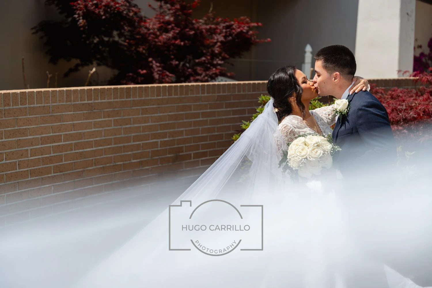 A bride and groom kiss outdoors, the bride holding a bouquet of white roses and wearing a lace wedding dress and veil, while the groom wears a dark blue suit with a white boutonniere. They stand in front of a brick wall with red and green foliage in 