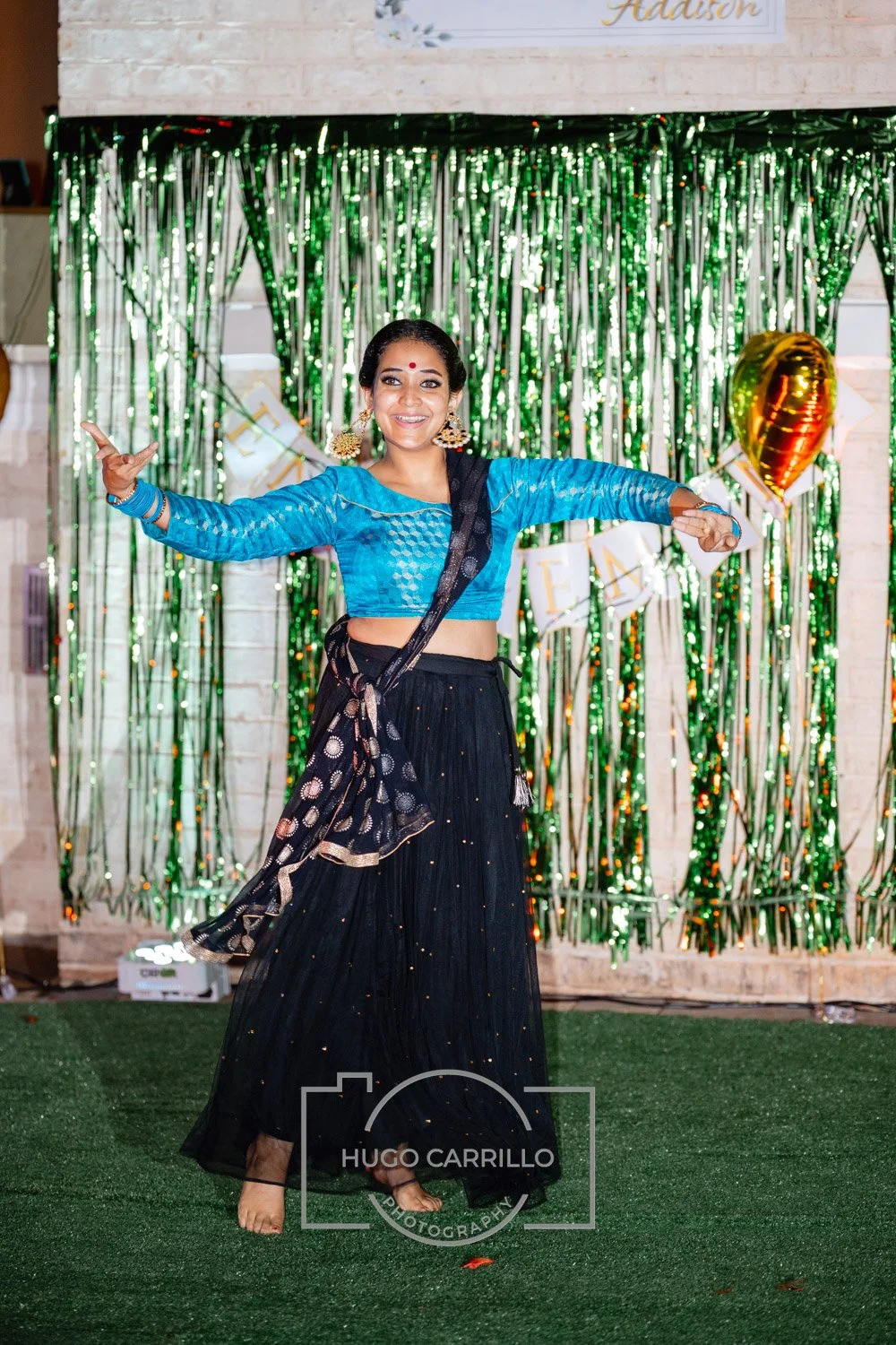 A woman dressed in traditional Indian attire dancing at a celebration event with green metallic streamers and balloons in the background.