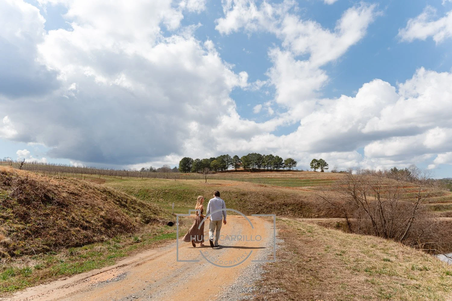 A couple walking together on a dirt path in a rural landscape with rolling hills, sparse trees, and a partly cloudy sky.