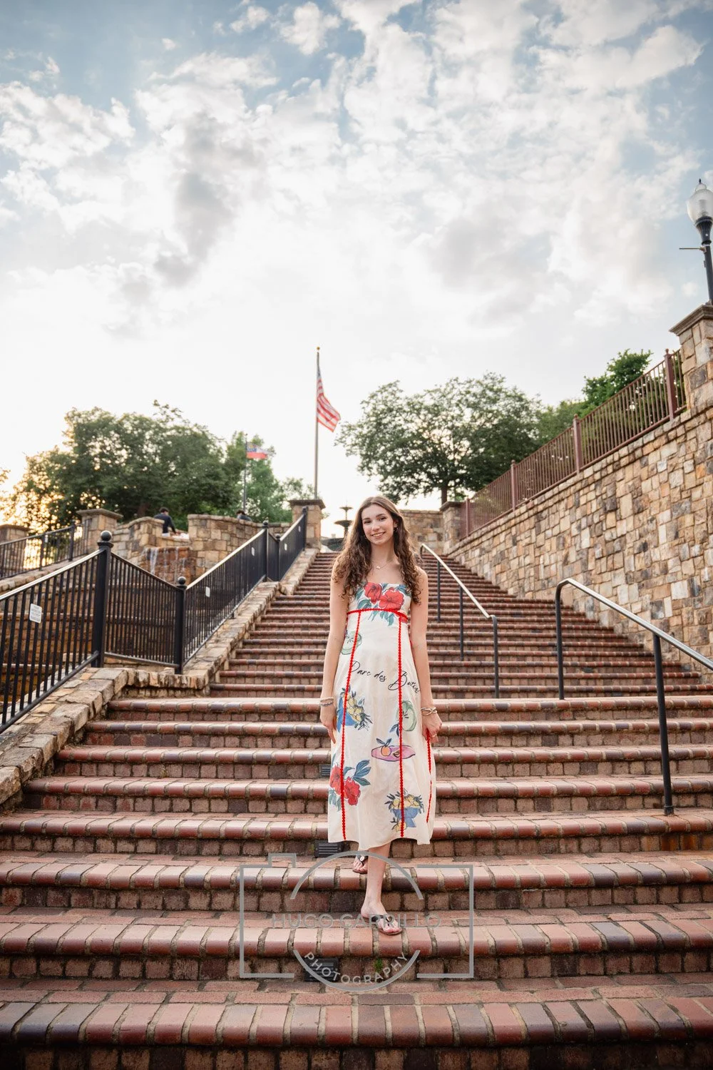 A woman in a colorful dress standing on brick stairs outdoors during sunset, with an American flag in the background.
