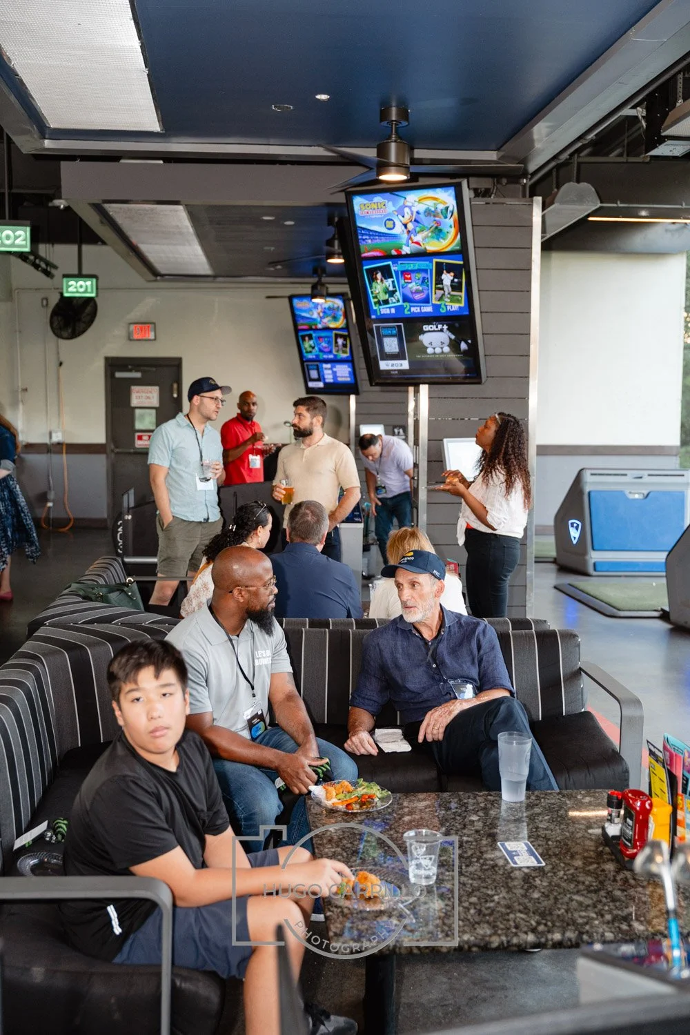 People socializing at a bar or lounge area, with large screens showing a sports game or entertainment, some seated and others standing with drinks in hand.
