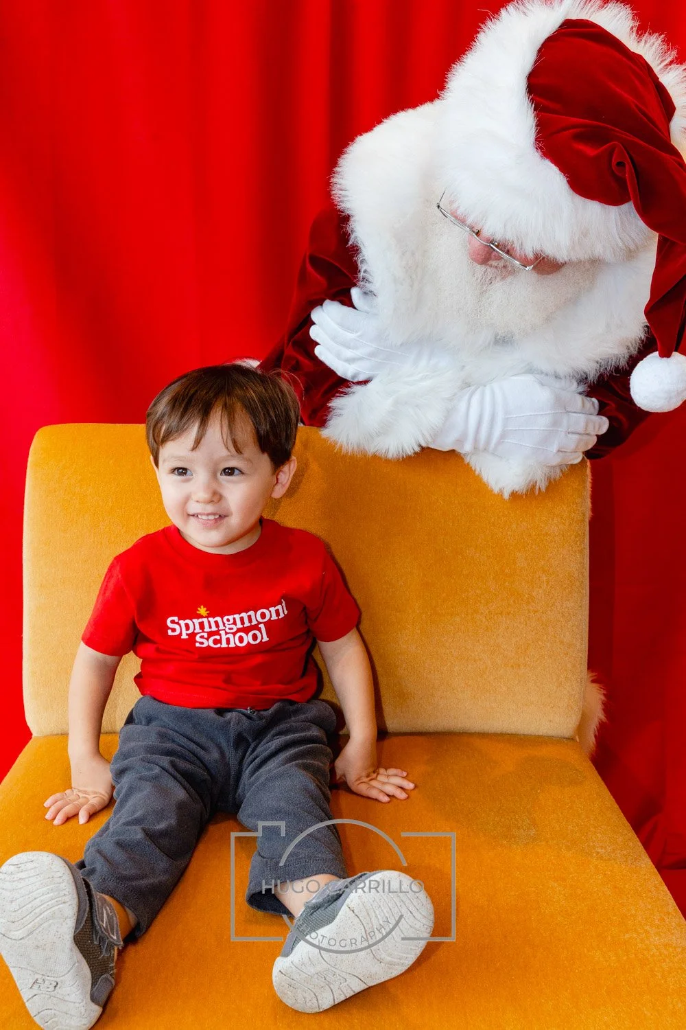 A young boy sitting on a yellow chair, smiling, wearing a red t-shirt with 'Springmont School' written on it, and gray pants. Behind him, a person dressed as Santa Claus leans over, with arms crossed, looking down at the boy, against a red backdrop.