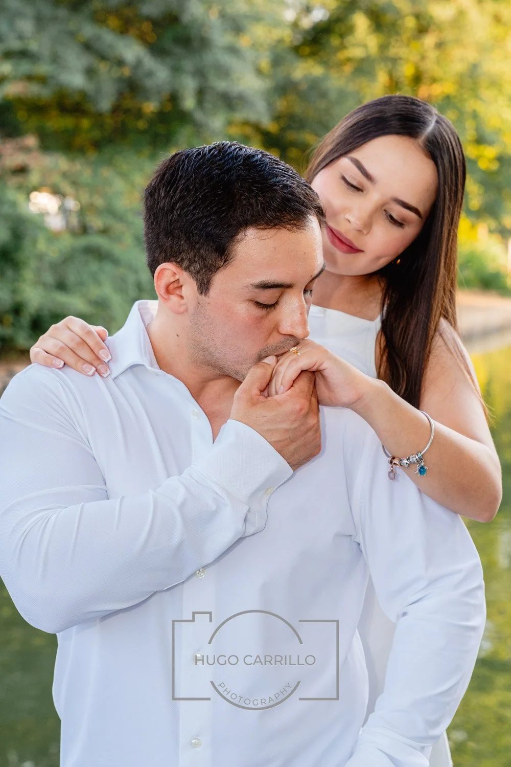 A man is kissing a woman's hand as he presses it to his lips. The woman is standing behind him, gently resting her hand on his shoulder, and smiling softly. They are outdoors near a body of water with green trees in the background.