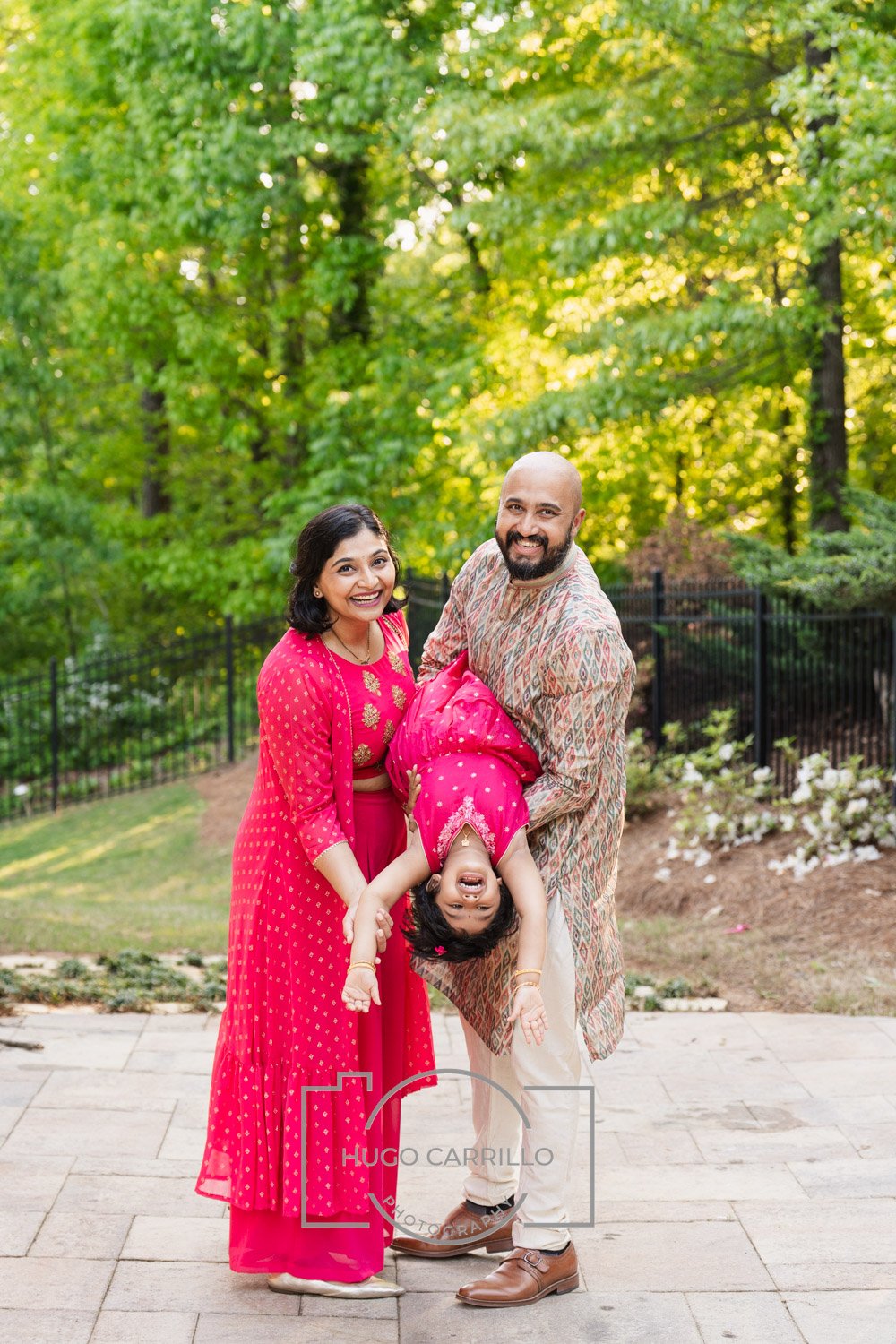 Family of three, of South Asian descent, outdoors in a park, smiling, with the father and mother holding a young girl upside down, wearing traditional Indian clothing, surrounded by greenery.