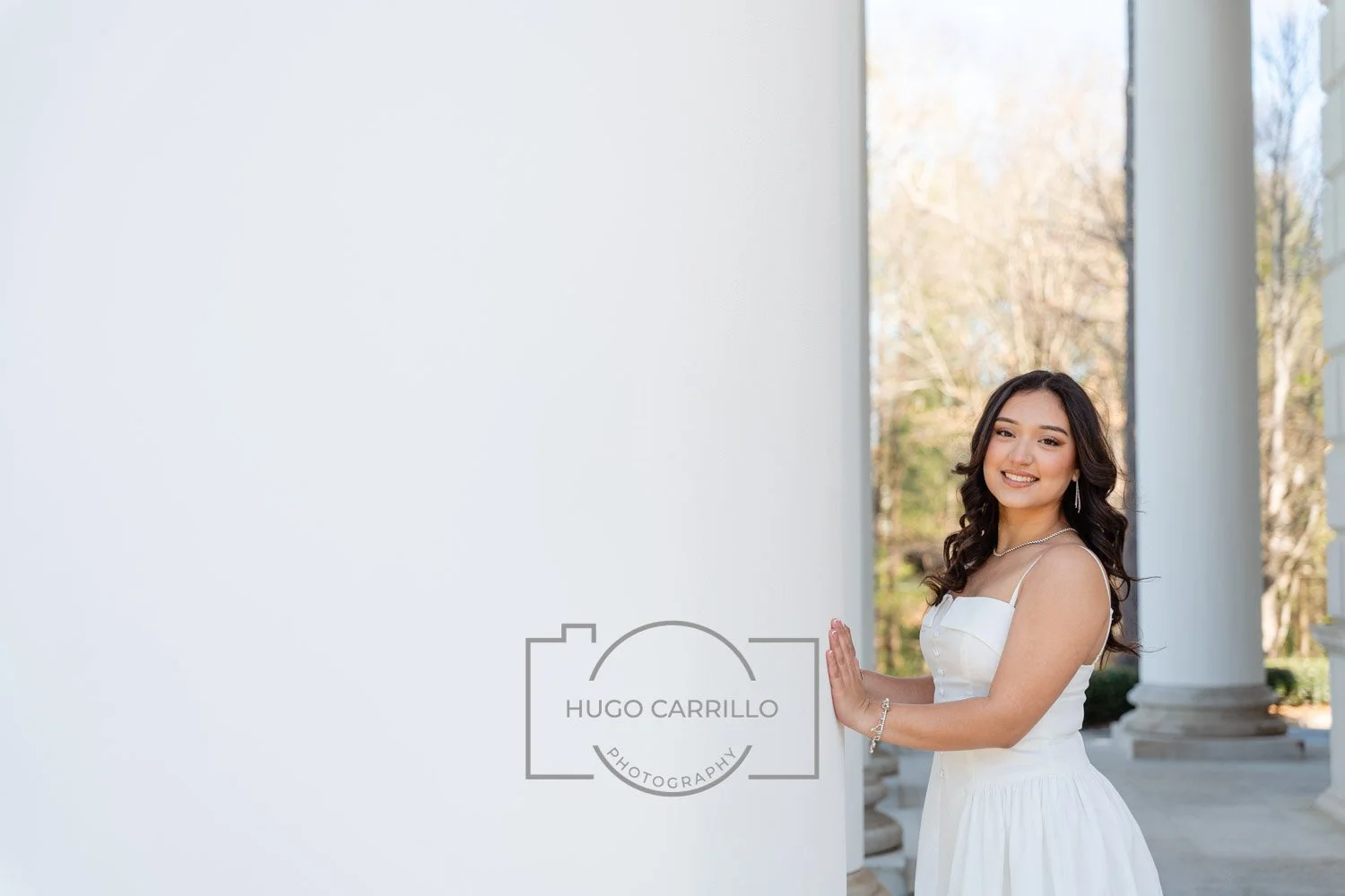 Young woman in white dress smiling and standing next to a white column outdoors with trees in the background.
