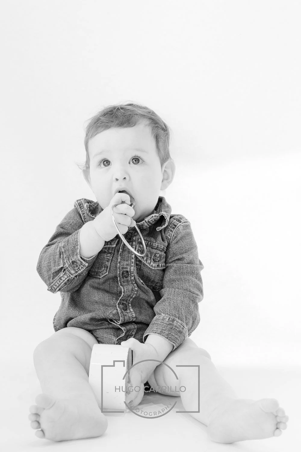 Black and white photo of a young child sitting on the floor, wearing a denim jacket, holding a toy phone, and with a surprised or curious expression.