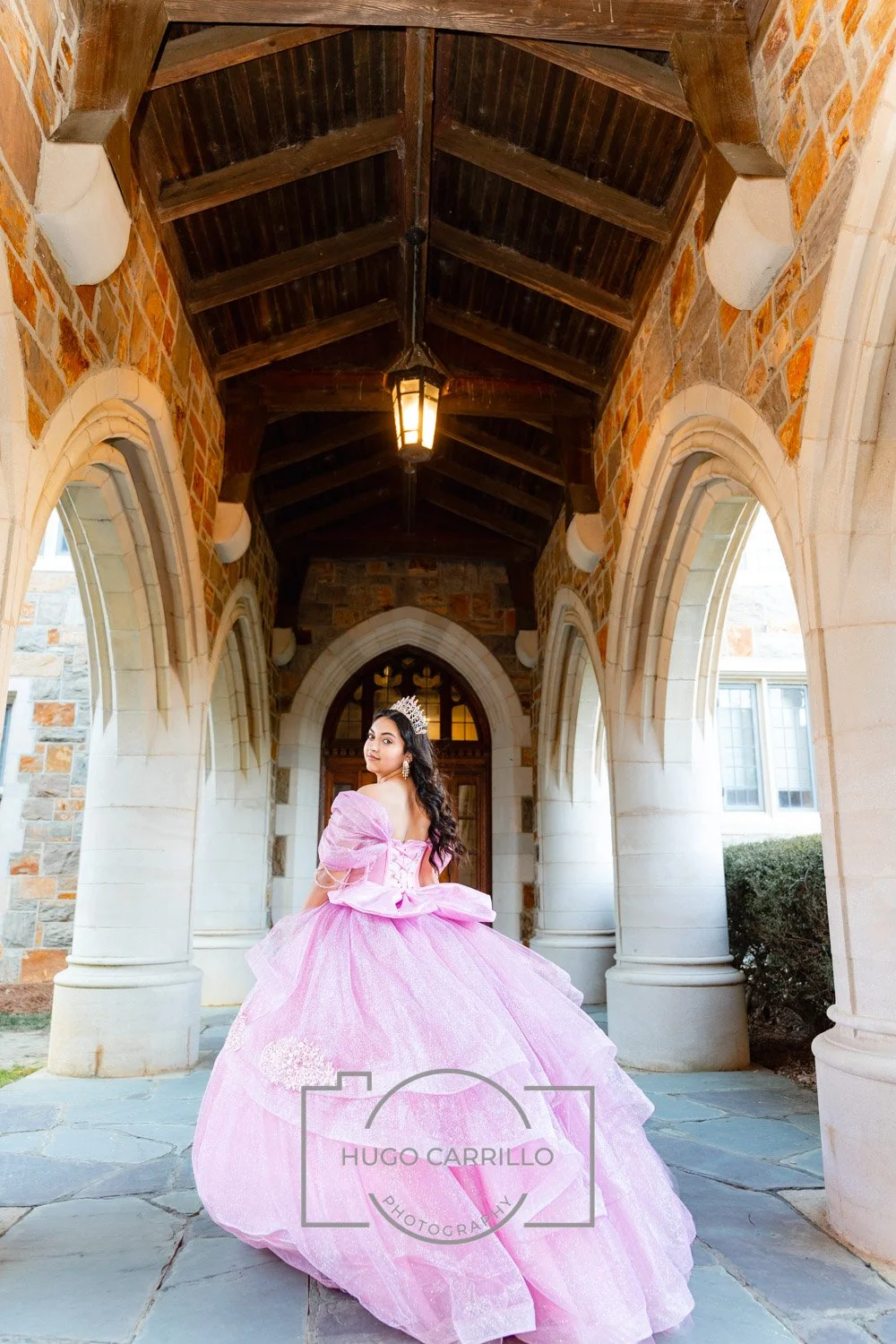 A quinceañera in a pink ball gown with off-the-shoulder sleeves and a tiara stands under a stone and brick archway in front of a wooden door, holding the skirt and looking over her shoulder.