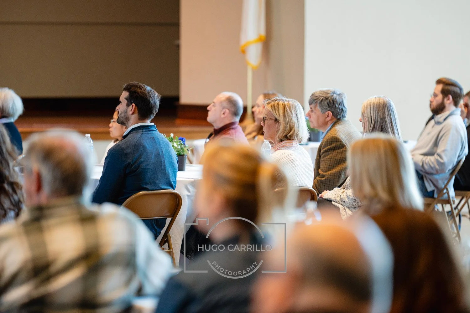 People attending a conference or seminar, sitting at tables in a large room, listening attentively.