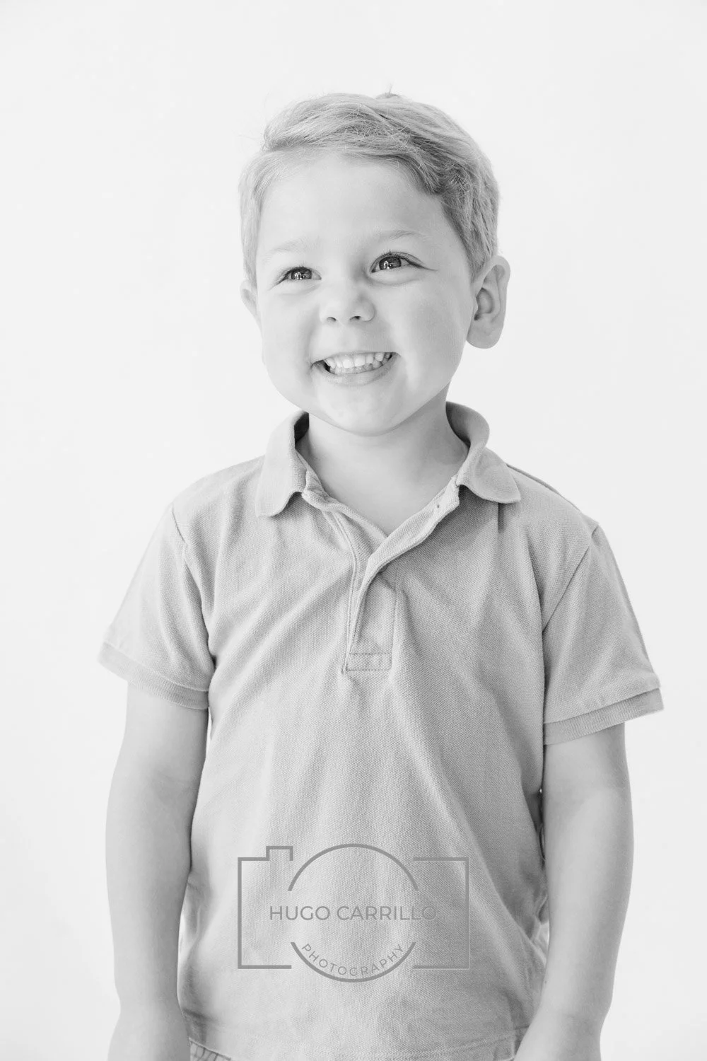 A smiling young boy with short hair wearing a polo shirt, standing against a plain background.