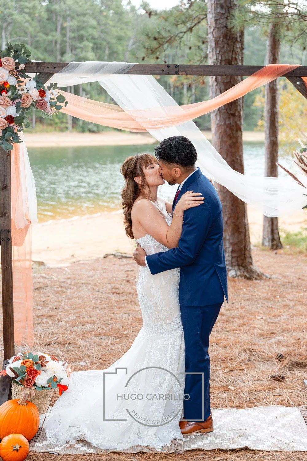 A bride and groom sharing a kiss outdoors under a decorated arch at a lakeside wedding with pumpkins and fall flowers.