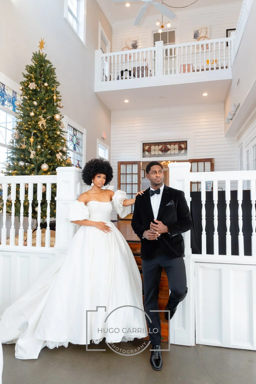 A bride in a white wedding gown with puffed sleeves and a groom in a black tuxedo with a bow tie stand inside a decorated home with a Christmas tree, white walls, and a high ceiling.