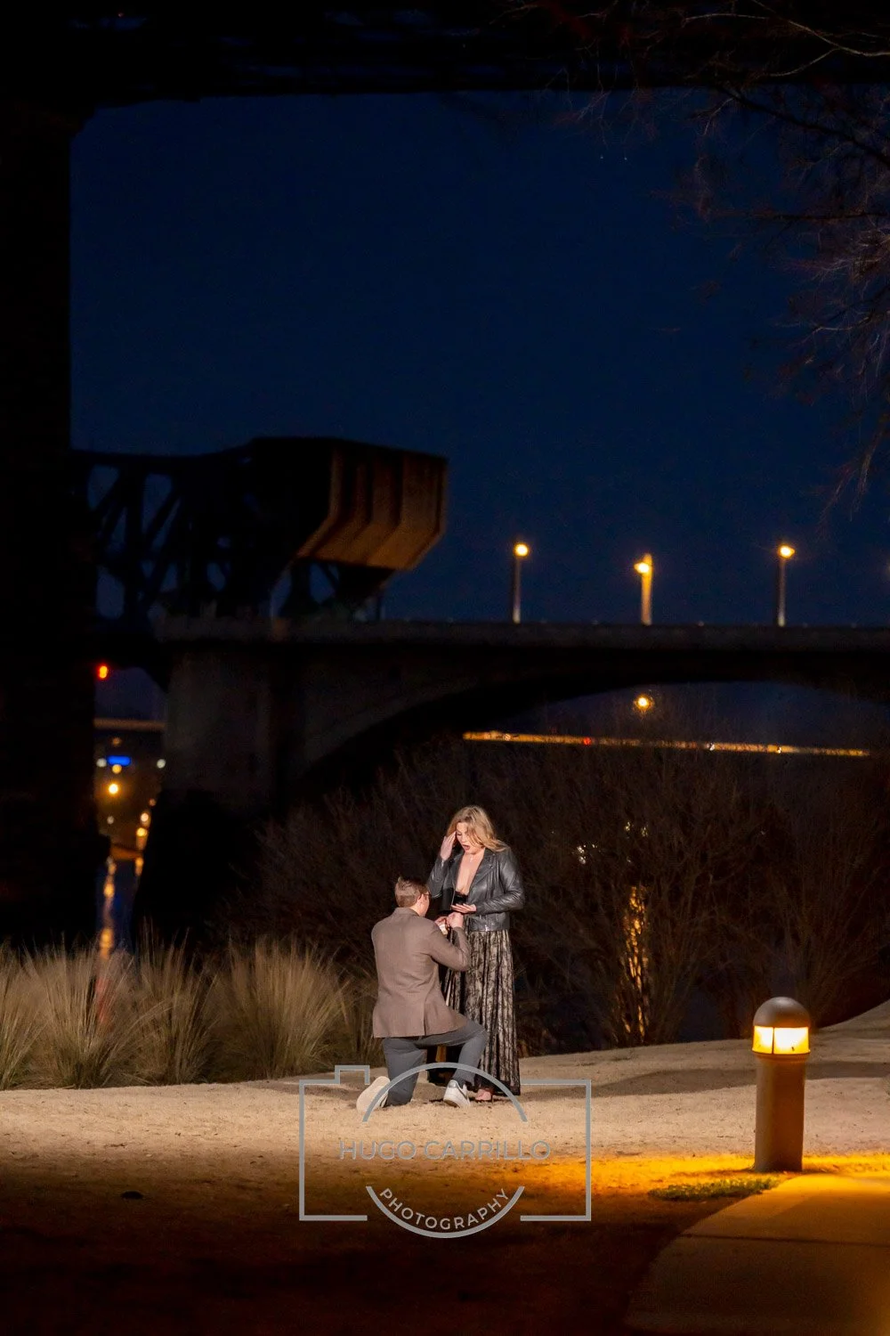 A man proposes marriage to a woman outdoors at night under streetlights, with a bridge in the background and a dark blue sky.