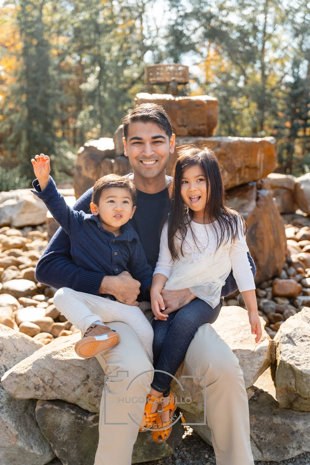 A man with two young children sitting on a large rock outdoors in front of a waterfall, surrounded by autumn trees.