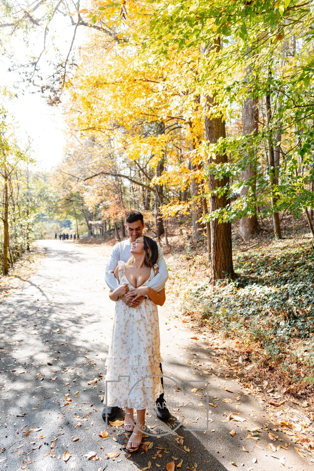 A couple embracing on a leaf-strewn path in a forest during autumn, with colorful fall foliage and sunlight filtering through the trees.