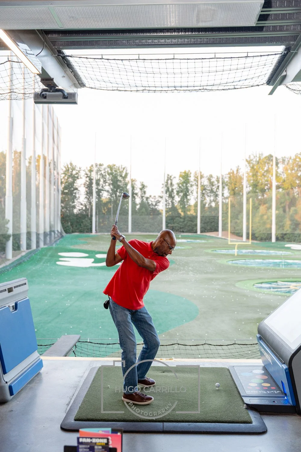 Man in red shirt and jeans practicing golf swing in indoor golf simulator with outdoor view in the background.