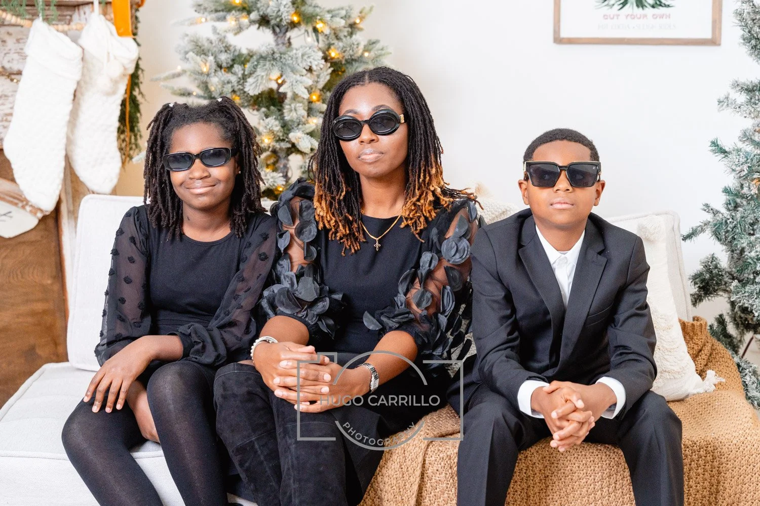 A woman and two kids sitting on a couch, all wearing black formal clothing and sunglasses, in front of a decorated Christmas tree with ornaments and stockings.