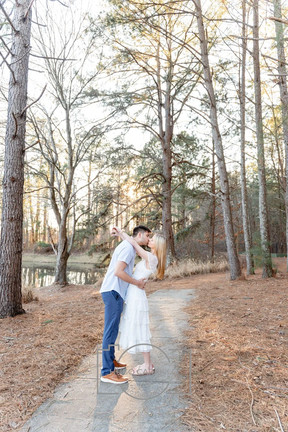 A couple sharing a kiss on a forest pathway near a lake at sunset, with tall trees around them.