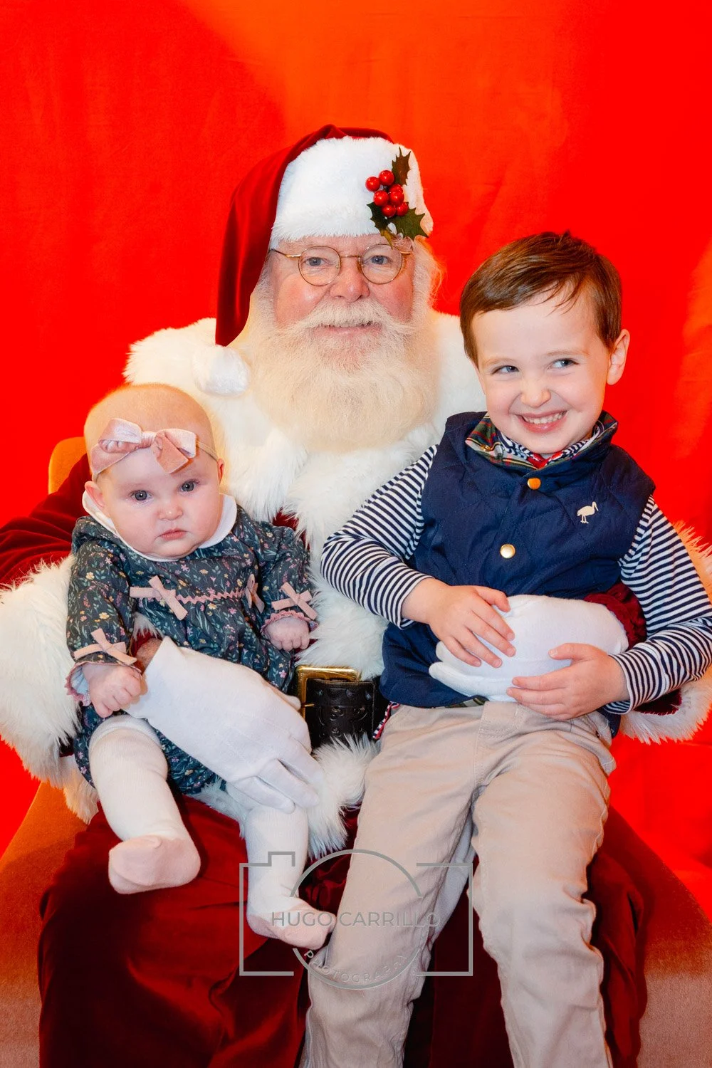 Two children sitting on Santa Claus's lap, a young boy smiling and a baby girl with a pink bow in her hair, against a red backdrop.