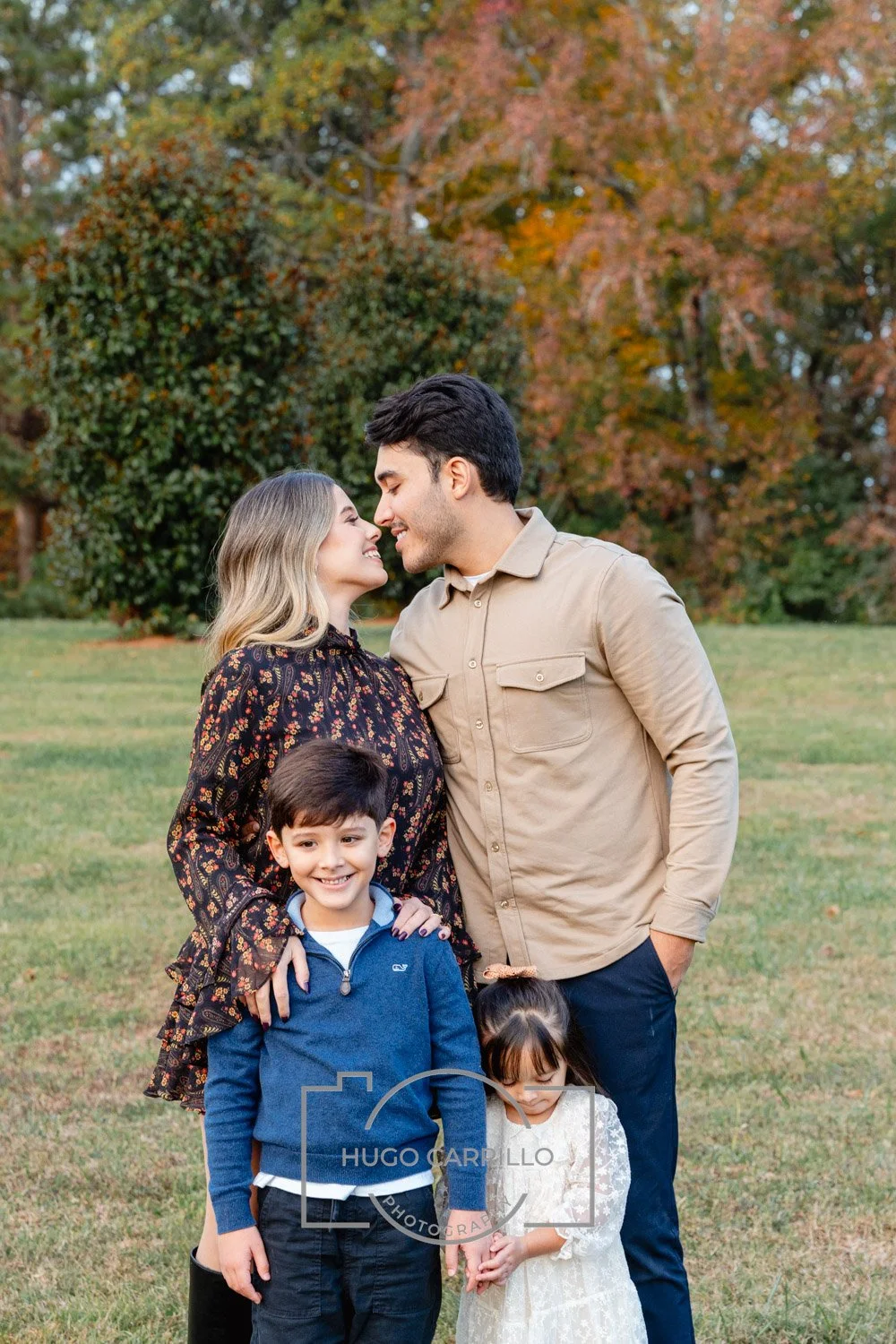 A family of four standing outdoors on a grassy area with autumn trees in the background. The parents, a man and woman, are smiling and leaning close together. Their two children, a boy and girl, stand in front of them, smiling and holding hands.