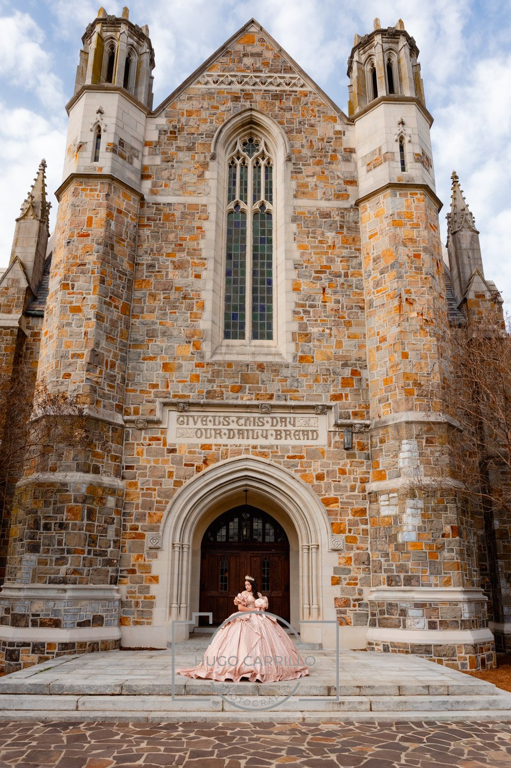 A quinceañera dressed in a pink ball gown with a tiara, standing on the steps of a stone church with Gothic architectural features, including pointed arches and tall stained glass windows.