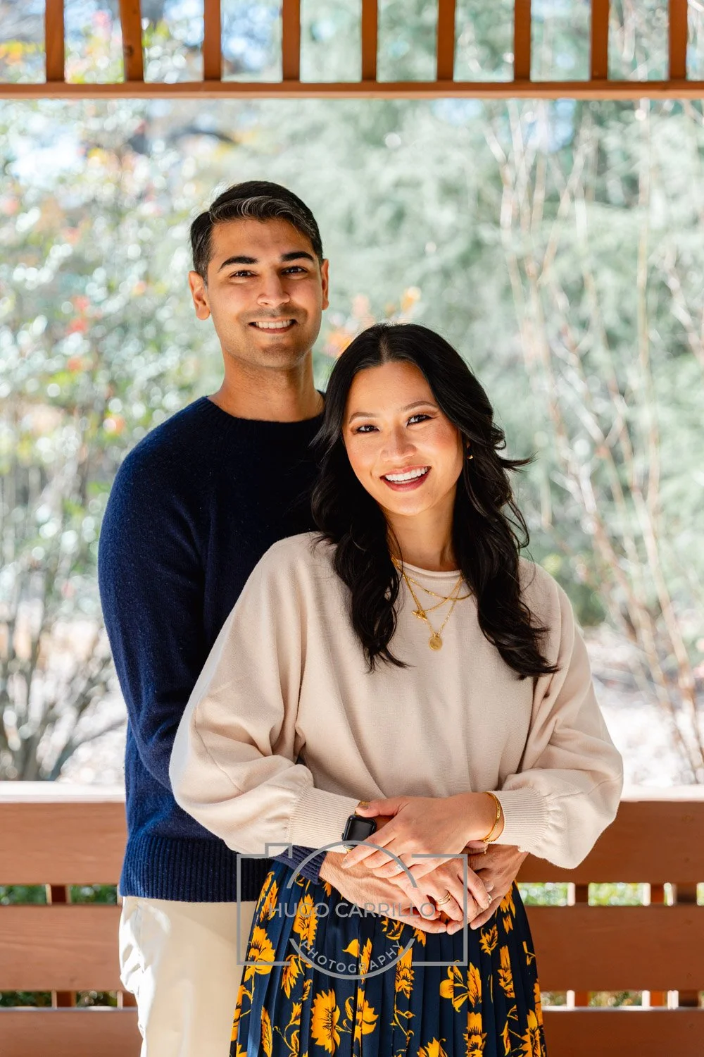 A smiling couple standing outdoors on a wooden porch, with trees in the background. The man is wearing a dark blue sweater and beige pants, and the woman is wearing a beige sweatshirt and a blue skirt with yellow floral patterns, accessorized with go