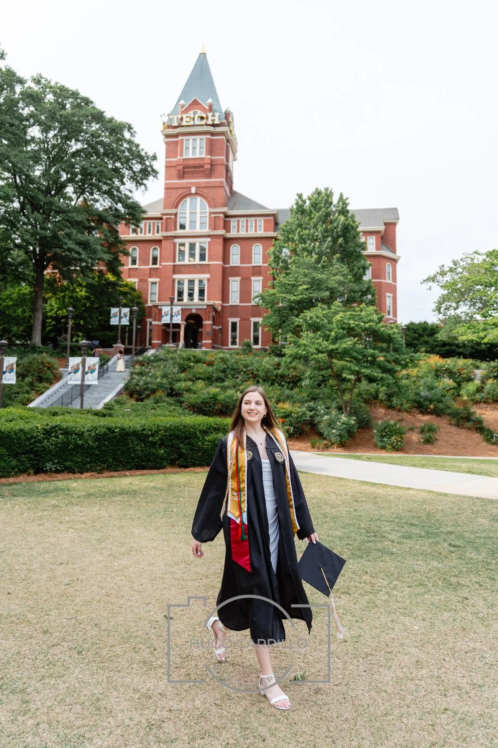 A young woman in a graduation gown and cap holding a diploma walking on a grassy area in front of a large, red brick building with a tower marked 'TECH'. She is smiling and surrounded by trees and landscaping.